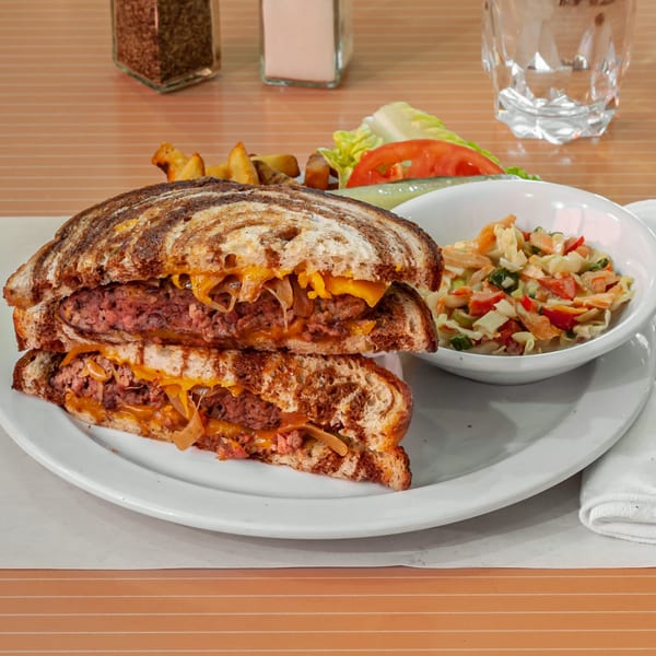 A patty melt with melted cheese and caramelized onions, served with coleslaw, fries, and a pickle.